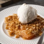 A serving of apple dump cake on a white plate topped with a scoop of ice cream, next to a spoon.