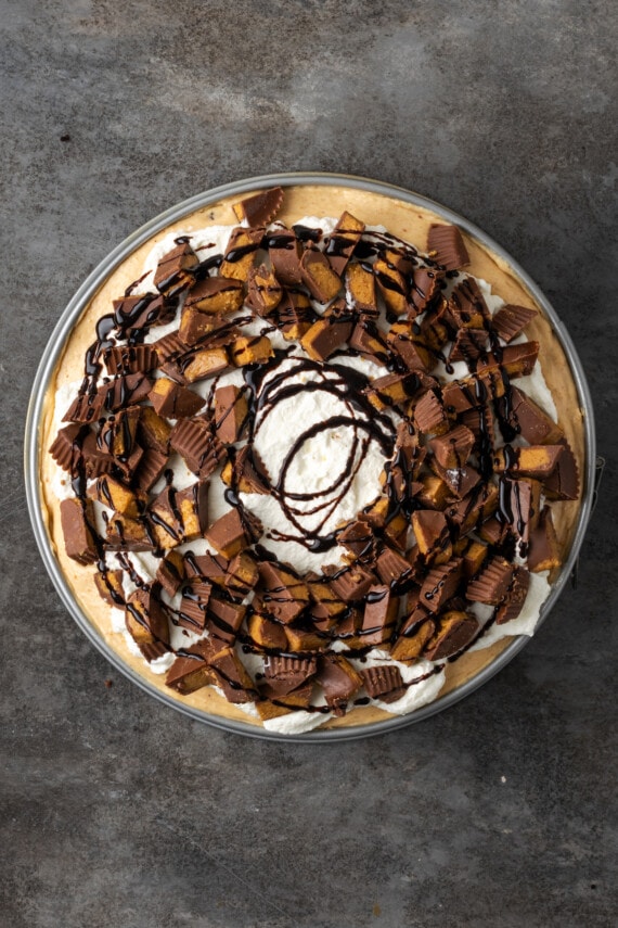 Overhead close up view of a finished brownie cheesecake topped with peanut butter cups and drizzled with fudge sauce on a white plate.