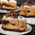 A slice of brownie cheesecake garnished with peanut butter cups on a white plate, with additional slices in the background.