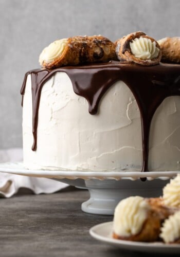 Cannoli cake on a cake stand drizzled with chocolate ganache and garnished with cannoli pastries.