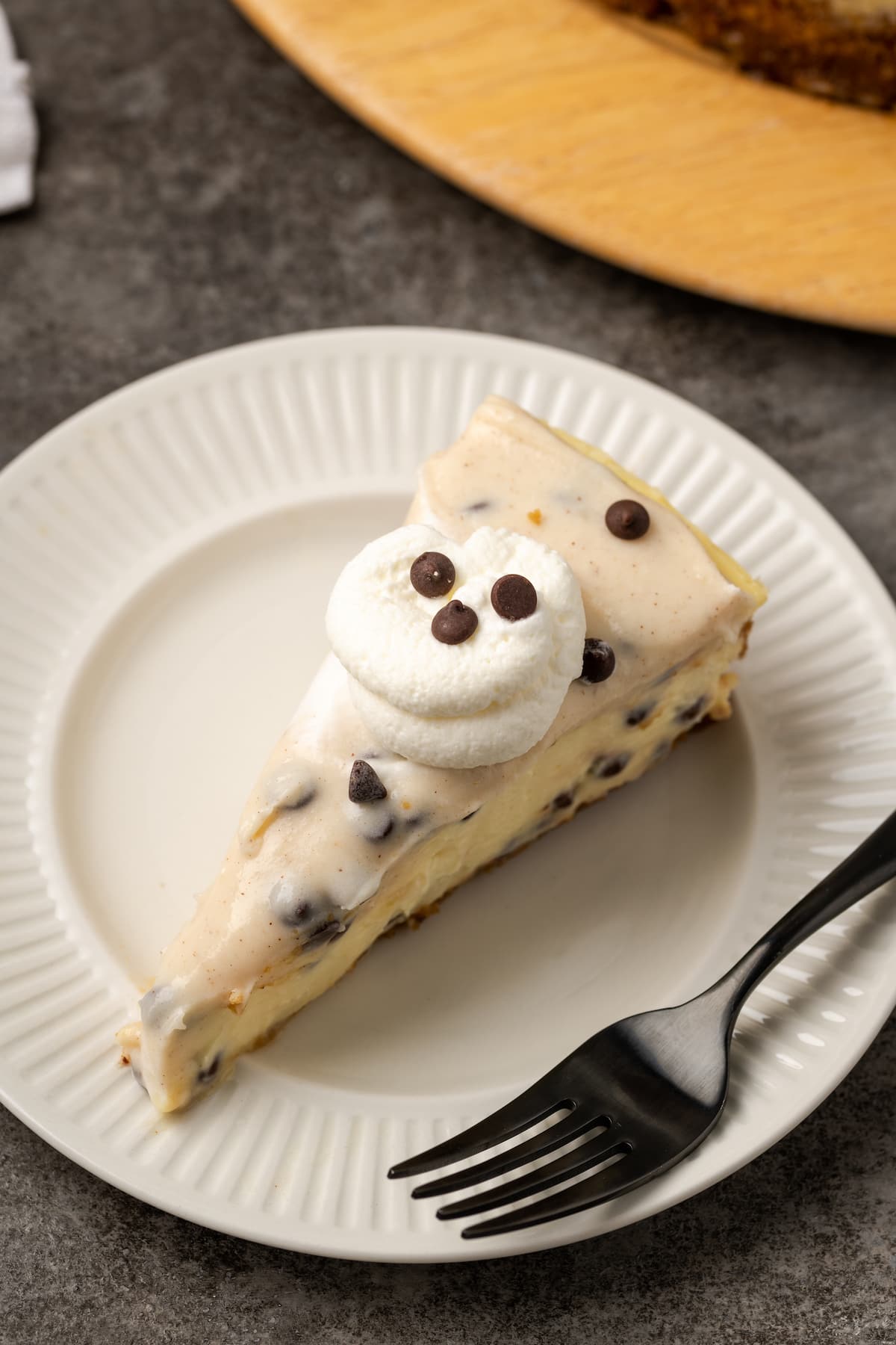 A slice of cannoli cheesecake topped with a swirl of whipped cream on a white plate next to a fork.