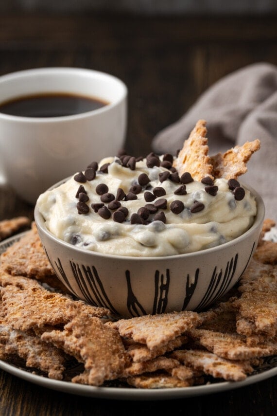 Cannoli dip served in a bowl with two cookies stuck into the dip, surrounded by more cookies on a plate with a cup of coffee in the background.