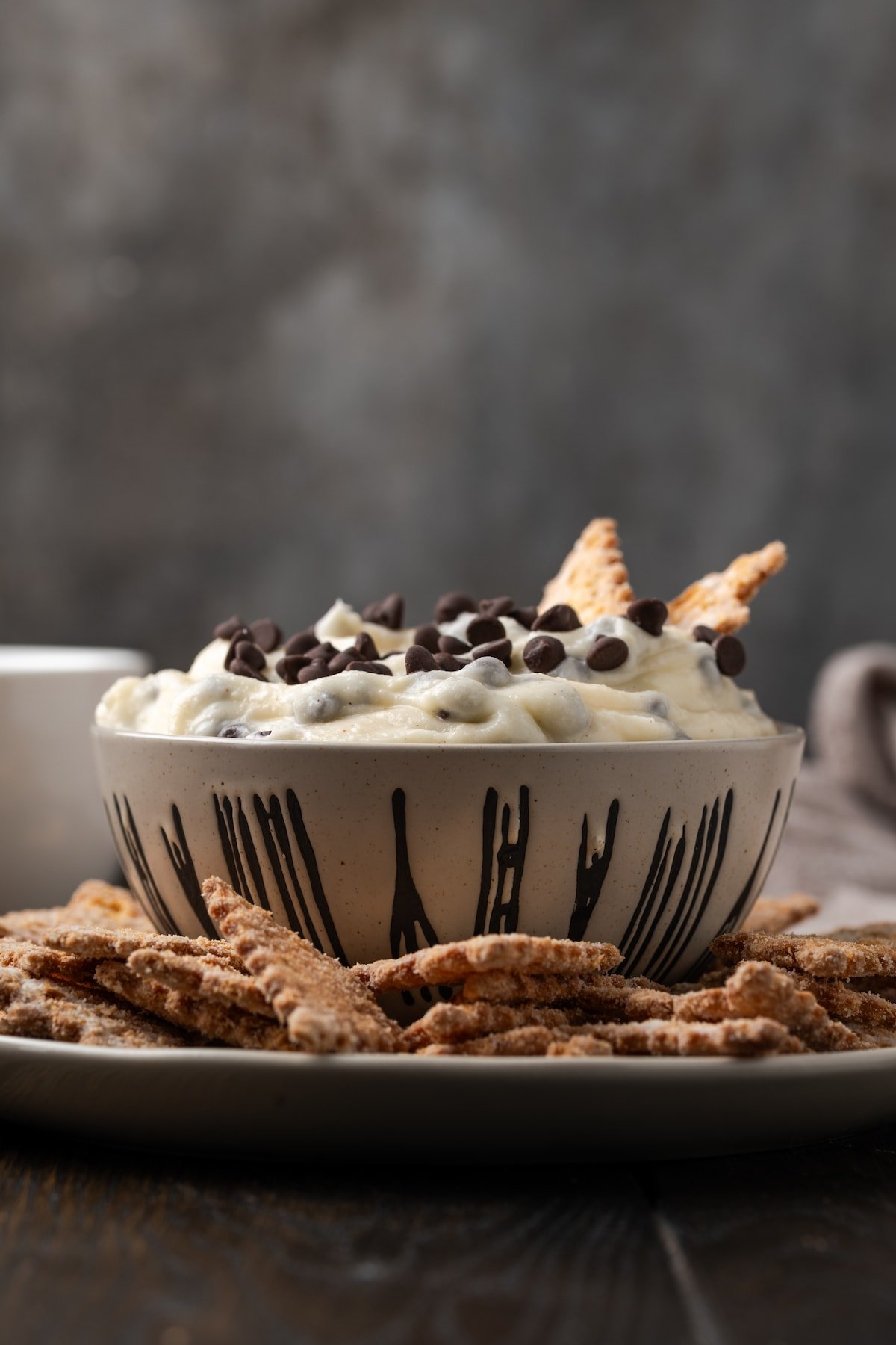 Cannoli dip served in a bowl with two cookies stuck into the dip, surrounded by more cookies on a plate with a cup of coffee in the background.