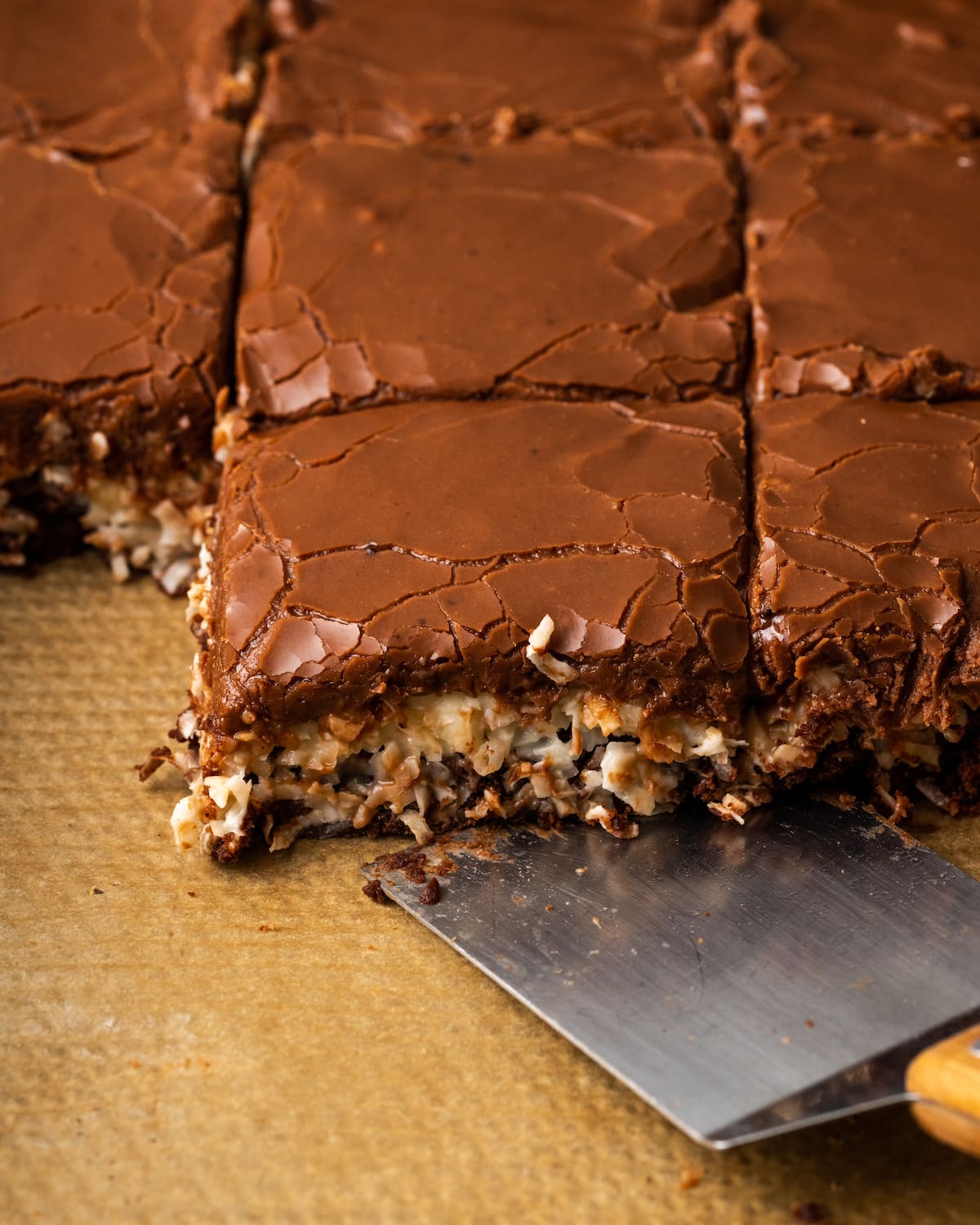 An offset spatula slides under coconut brownies in a lined pan.