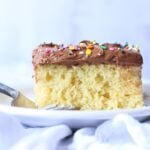 Vanilla cake with chocolate frosting and rainbow sprinkles on a white plate with a fork.