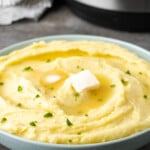 Creamy Instant Pot mashed potatoes in a bowl topped with two pats of butter and chives, with the Instant Pot in the background.