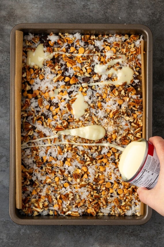 A hand pours a can of sweetened condensed milk over magic bar ingredients in a lined baking pan.