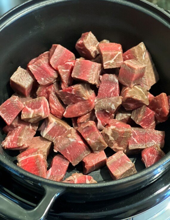 A Crockpot insert with cubes of raw sirloin steak in the bottom.