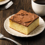 A slice of frosted sour cream coffee cake on a white plate next to a fork and a cup of coffee.