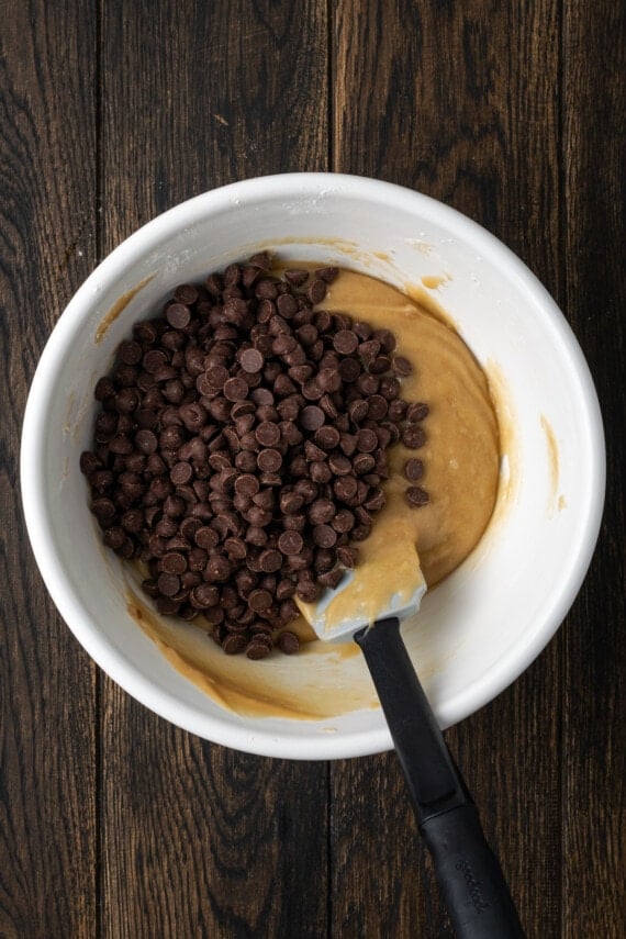 Chocolate chips added to cookie batter in a white bowl, with a spatula.