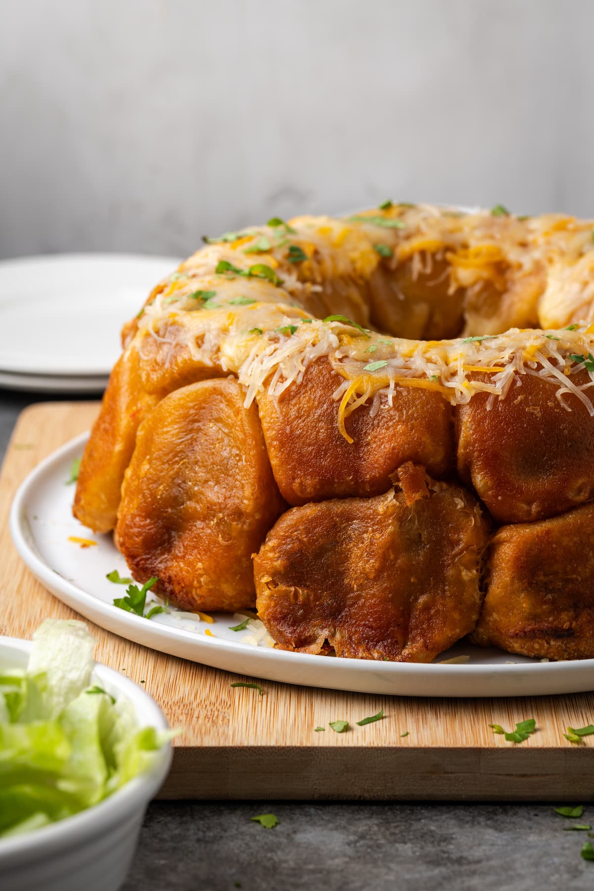 Taco monkey bread topped with melted cheese on a white plate, next to a bowl of shredded lettuce.