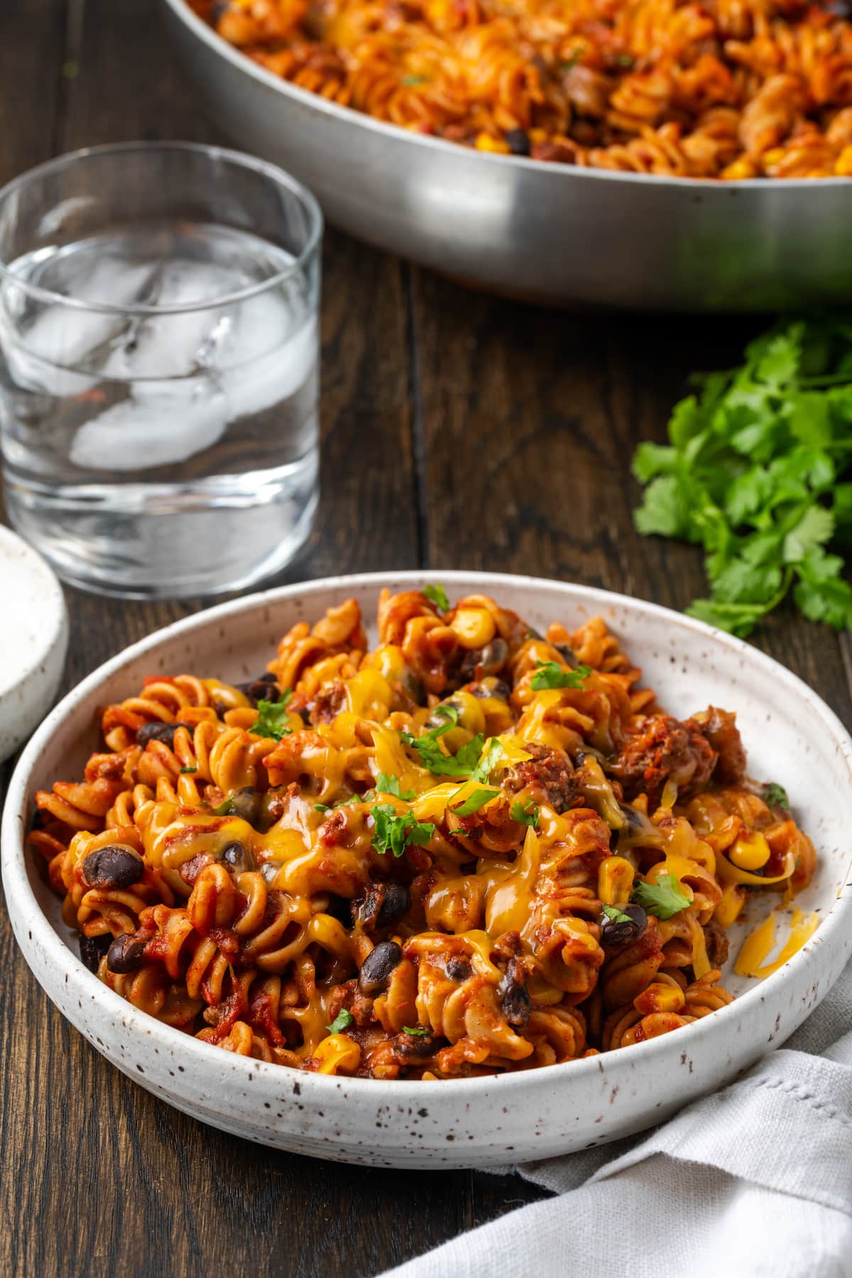 Taco pasta in a white bowl next to a fork, with a glass of ice water and a skillet of taco pasta in the background.